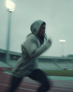 A runner in a hoodie sprints on a track under stadium lights, captured with motion blur for a dynamic and intense feel.