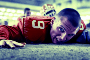 Football player in a red uniform lying face-down on the field after a tackle, showing determination and intensity.