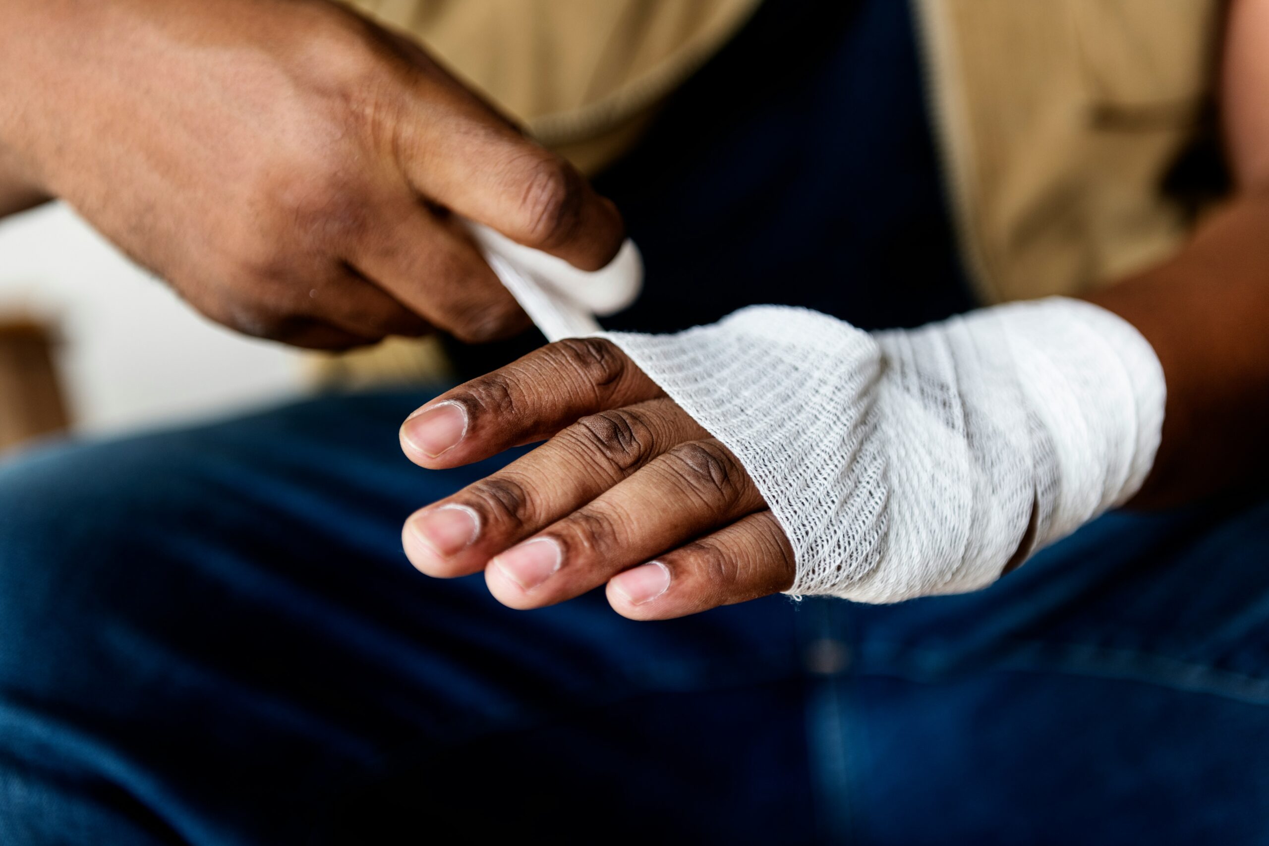 Close-up of a person’s hands as one hand carefully wraps a white gauze bandage around the other hand, which is partially covered and supported, with fingers exposed and resting over dark blue jeans, showing the texture of the bandage and the careful motion of securing it in place.