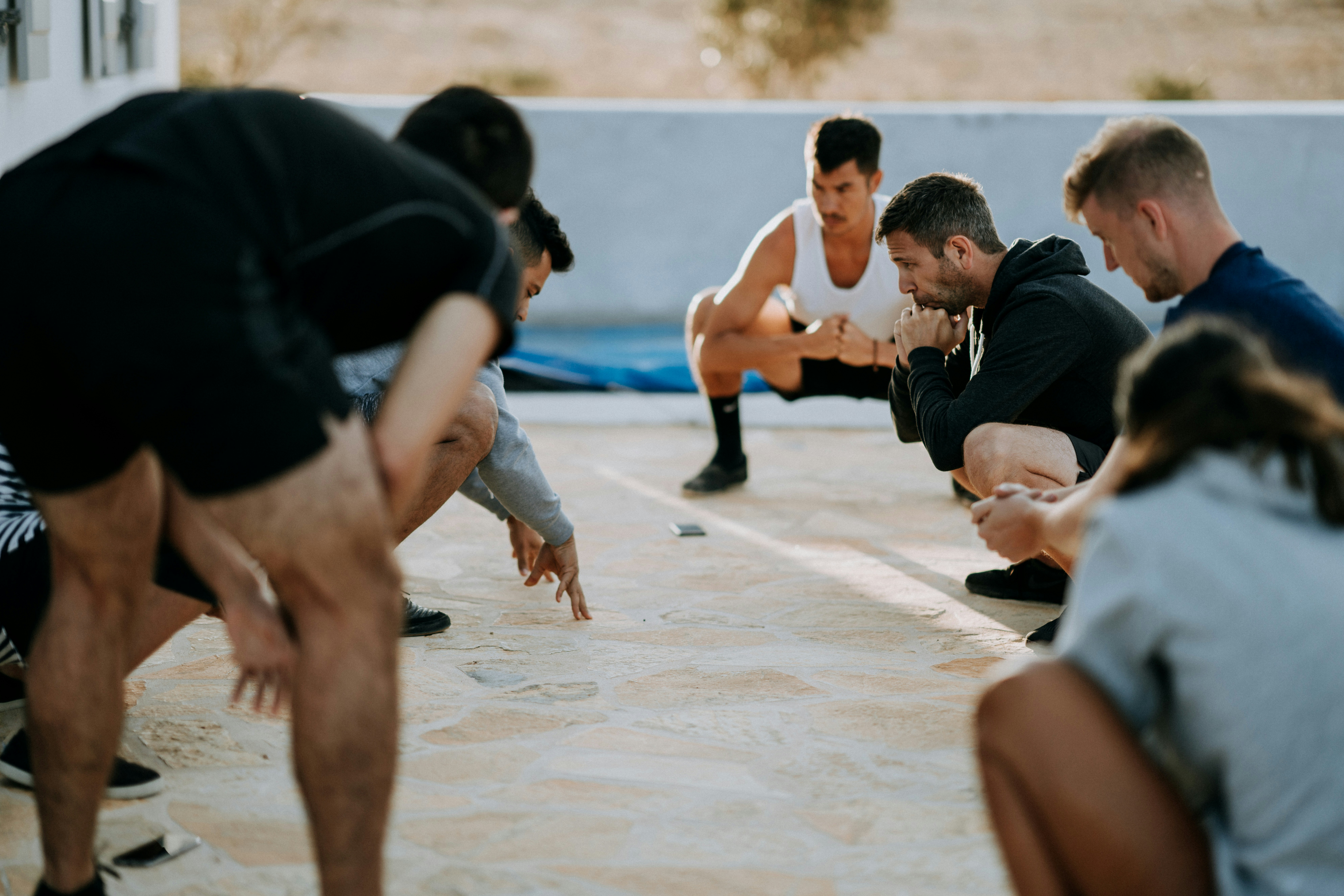 A group of athletic adults crouch in a tight circle on a sunlit outdoor patio, focusing intently on the ground as if preparing for a group exercise or strategy drill, with muscular legs, workout clothing, and concentrated expressions suggesting a high-intensity training session.