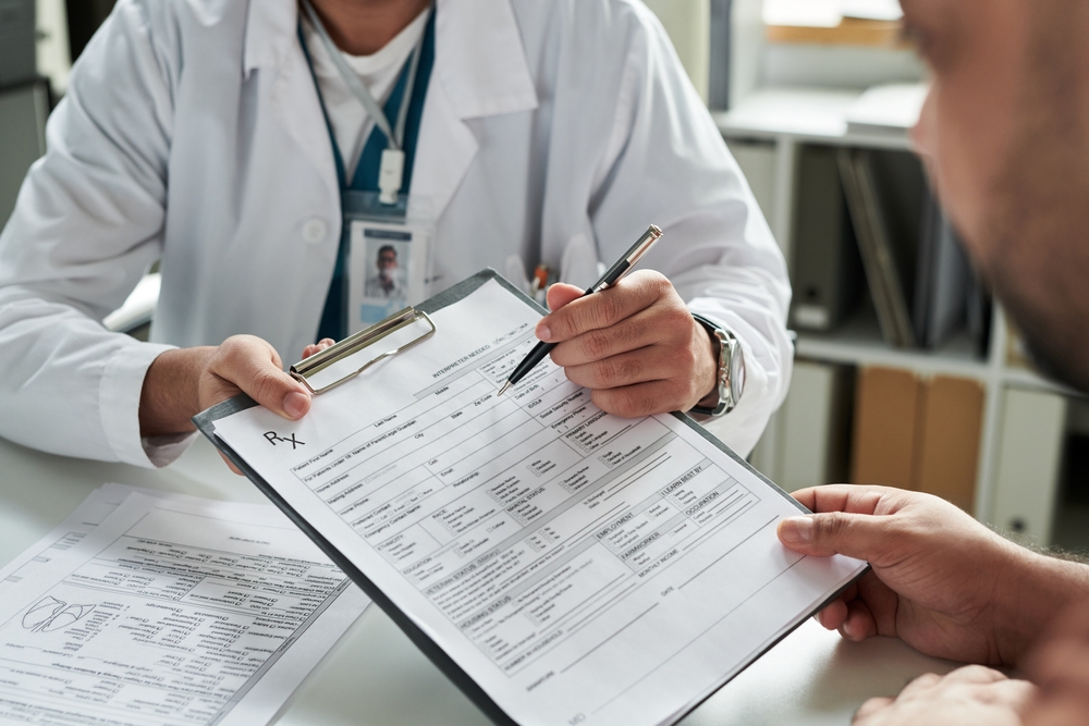 A doctor writes on a clipboard while a man observes attentively beside him.